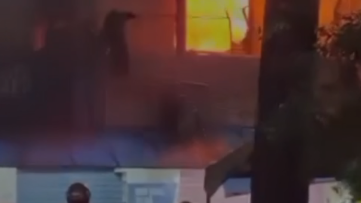 Cena noturna com iluminação vermelha e fumaça sugerindo incêndio em residência no Guarujá, com pessoas na área ao fundo e marco de janela ao centro