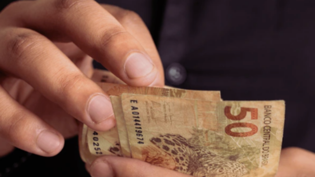Quinto dia útil de agosto 2026: Mãos segurando cédula de dinheiro de R$50 em contexto de pagamento de salários; texto ao fundo indica prazo para empresas realizarem o pagamento de julho. (Foto: Canva)