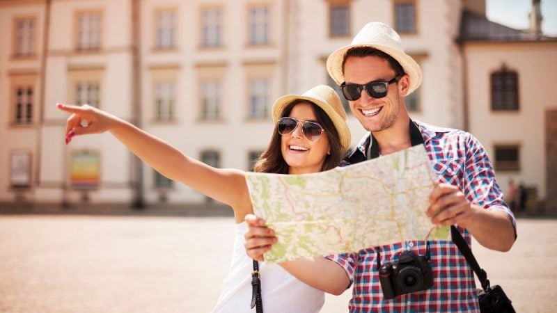 Casal de turistas com chapéus e mapa, em viagem turística no centro de uma cidade, destacando faturamento do turismo com movimentação de R$ 23,2 bilhões em maio de 2026.