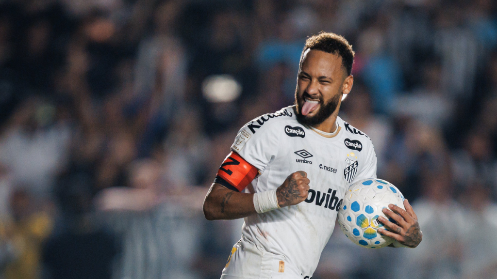 Neymar celebra gol pelo Santos em partida no estádio, segurando a bola e comemorando com expressão confiante; Foto: Raul Baretta / Santos FC.