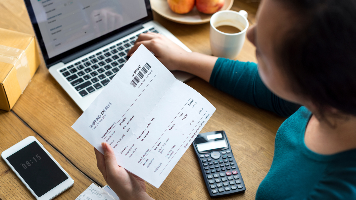 Pessoa analisa uma nota fiscal em uma mesa com calculadora e notebook ao fundo, destacando o início de novos impostos da Reforma Tributária nas compras.