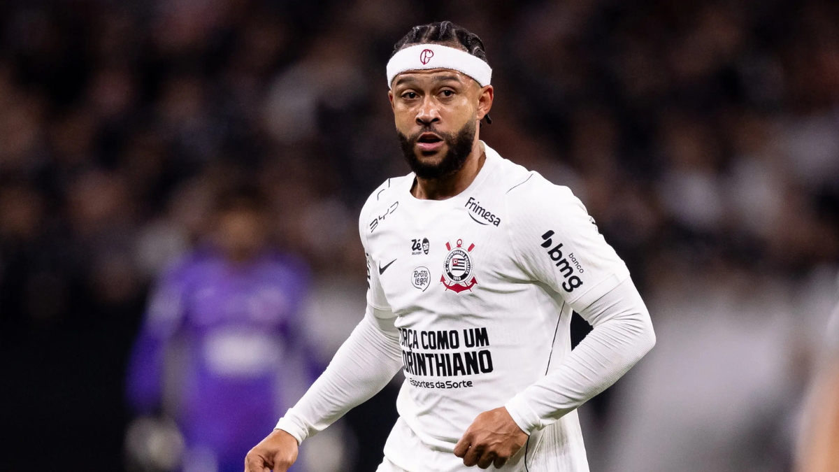 Memphis é retratado em campo com camisa branca do Corinthians durante partida, ao fundo há torcedores desfocados no estádio e o jogador posa em postura de jogo.