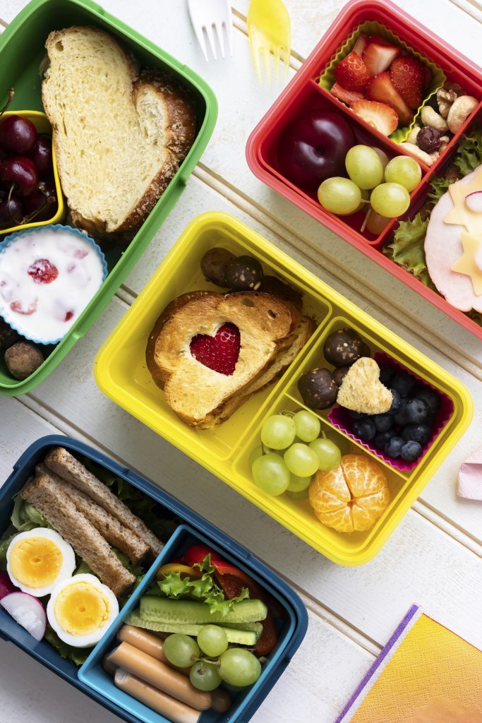 Lanches saudáveis em potes coloridos para crianças, com fatias de pão, frutas como uvas e morangos, além de iogurte, granola e palitinhos em marmitinhas