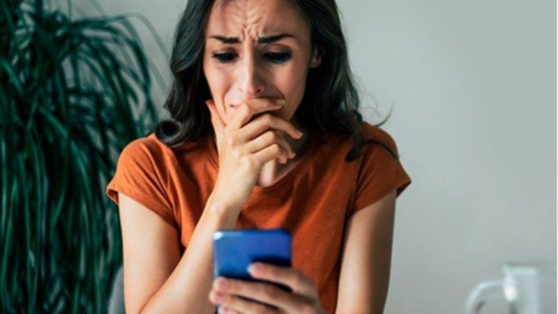 Mulher com expressão preocupada, segurando um celular na sala de estar, em cena que destaca a necessidade de medida protetiva lei maria da penha para transformar proteção em segurança real após anos de desafios.