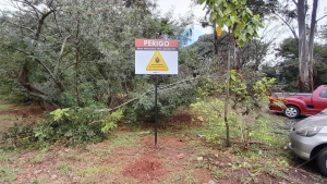 Placa de aviso de perigo em área de risco próximo a vegetação e carros estacionados, em cenário de investigação ligado à febre maculosa e mortes confirmadas.