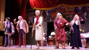 Musical Forever Young em cena: atores encenam com figurinos coloridos em um palco, com cenário temático ao fundo e iluminação de teatro. Carmo Dalla Vecchia e Vanessa Gerbelli se apresentam.