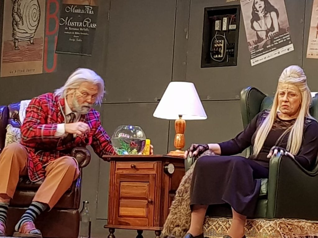 Musical Forever Young: Cena de comédia musical com dois atores caracterizados como personagens idosos sentados em cadeiras, em palco, divulgando apresentações nos dias 13 e 14 de agosto. Divulgação Estevan Buzzato