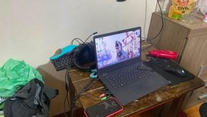 Mesa com notebook aberto mostrando vídeo e documentos, teclado, mouse e fones; ao lado há celular, cabos e objetos pessoais, em cenário que remete a investigação sobre Eleições 2026 e Grupo extremista.