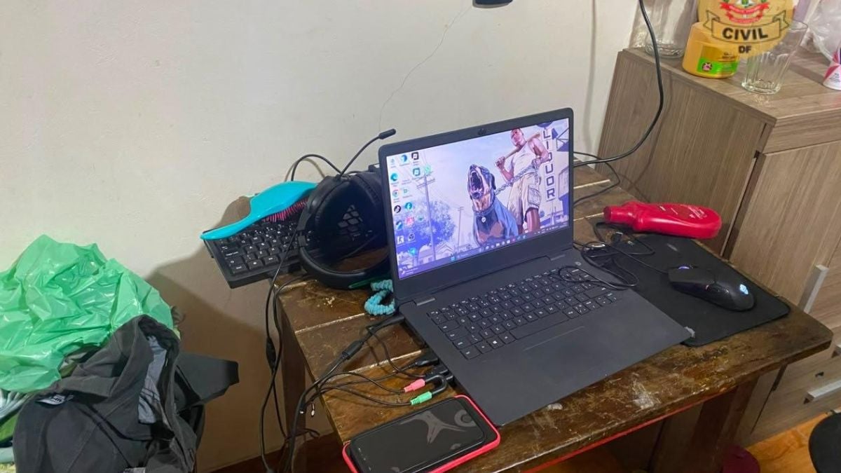 Mesa com notebook aberto mostrando vídeo e documentos, teclado, mouse e fones; ao lado há celular, cabos e objetos pessoais, em cenário que remete a investigação sobre Eleições 2026 e Grupo extremista.