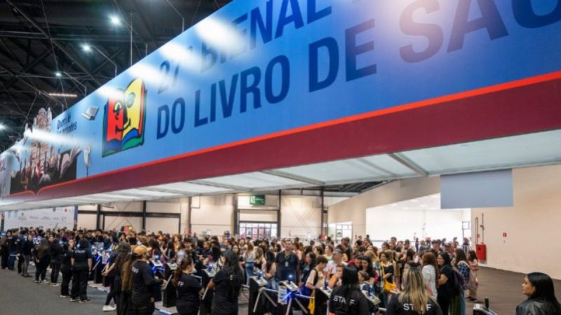 Bienal do Livro terá maior edição da história e ingresso com cashback total em foto de público reunido em área de estande, com banner “Dia do Livro” ao fundo em evento.