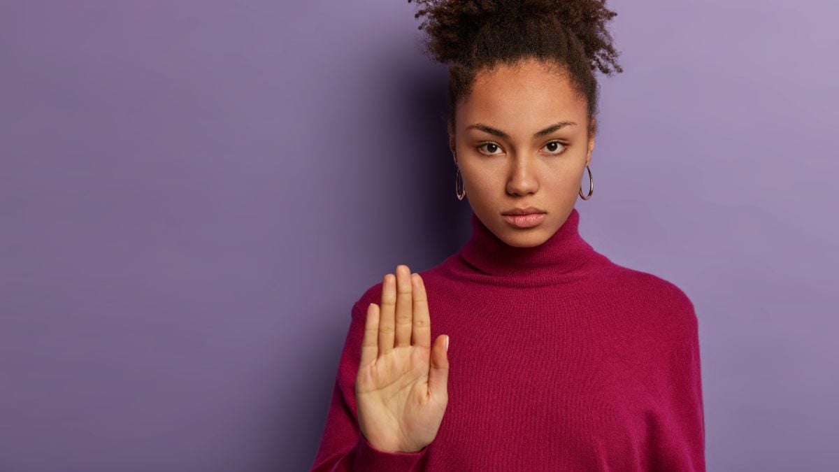 briga em loja de informática: Mulher com expressão séria e postura firme, mantendo a mão levantada em sinal de “pare”, em cenário com fundo roxo, em contexto de violência contra a mulher.