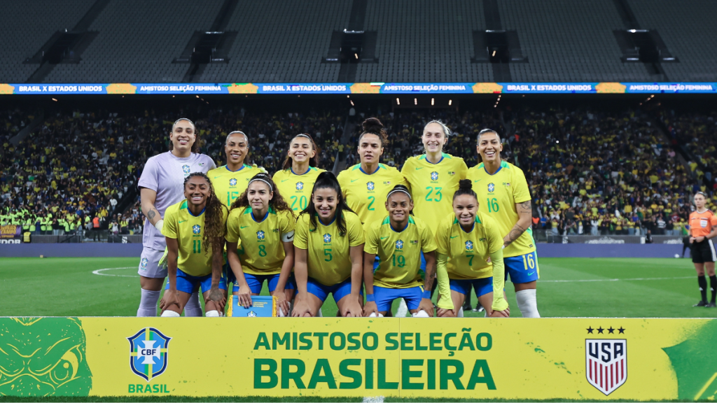 Seleção Brasileira Feminina em foto de equipe no estádio, com jogadoras uniformizadas durante amistoso; banner “Amistoso Seleção Brasileira” e em fundo “Copa do Mundo Feminina de 2027”.