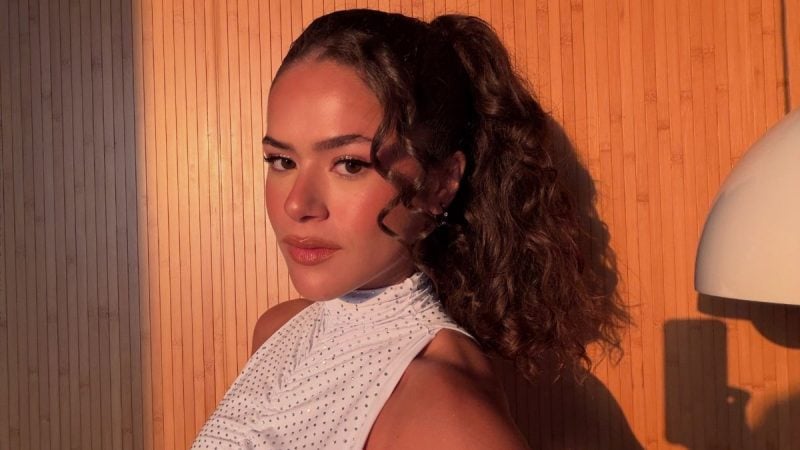 Maisa em foto de bastidores, com cabelo preso e vestido branco, em ambiente com iluminação quente e fundo laranja, durante entrevista sobre crítica e régua