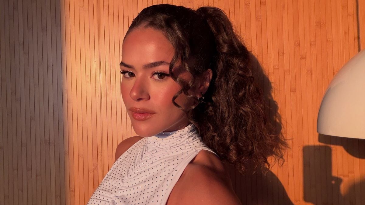 Maisa em foto de bastidores, com cabelo preso e vestido branco, em ambiente com iluminação quente e fundo laranja, durante entrevista sobre crítica e régua