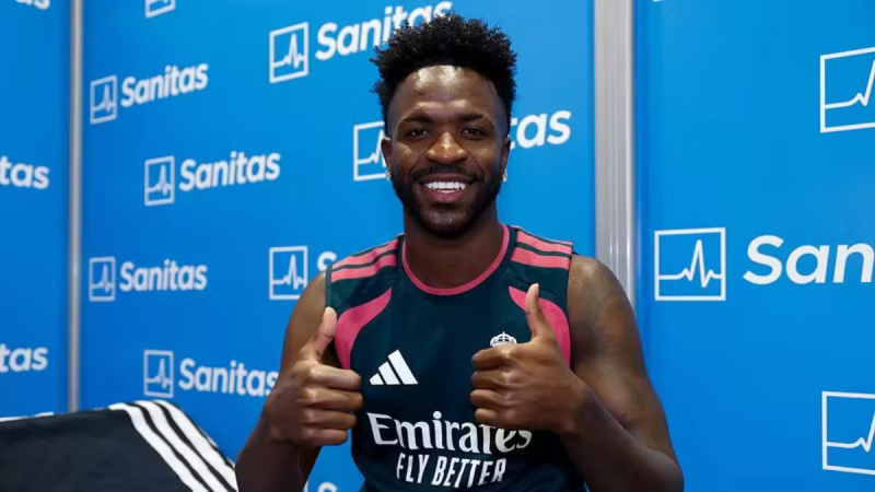 Vini Jr sorri em coletiva de imprensa no Real Madrid, em frente a um painel azul com a marca Sanitas, com as mãos fazendo sinal de positivo.