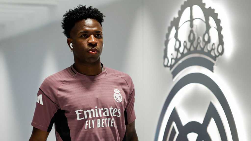 Vini Jr. em foto com uniforme do Real Madrid, em ambiente interno, ao lado do escudo do clube; imagem de reprodução do X @realmadrid.
