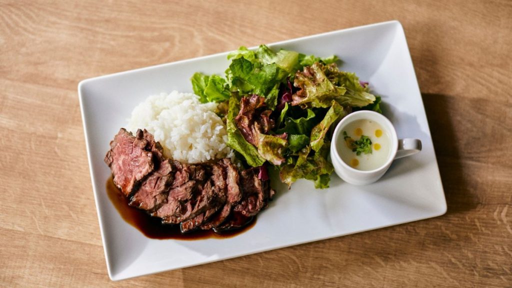 Prato retangular com porção de arroz branco, carne assada e salada verde ao lado de um molho em uma mesa de madeira, retratando consumo de arroz no Brasil.