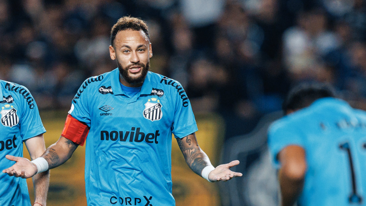 Neymar em partida de futebol, com uniforme do Santos, gesticulando com as mãos no campo. Imagem de lance próximo da torcida ao fundo.