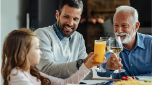 Família reunida à mesa em restaurante durante o Dia dos Pais, com pai idoso e filho brindando com sucos e sorriso, em clima de comemoração