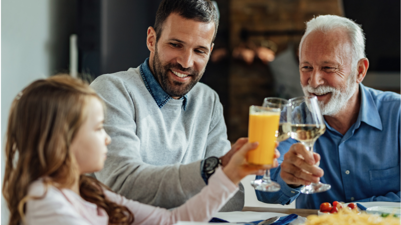 Família reunida à mesa em restaurante durante o Dia dos Pais, com pai idoso e filho brindando com sucos e sorriso, em clima de comemoração
