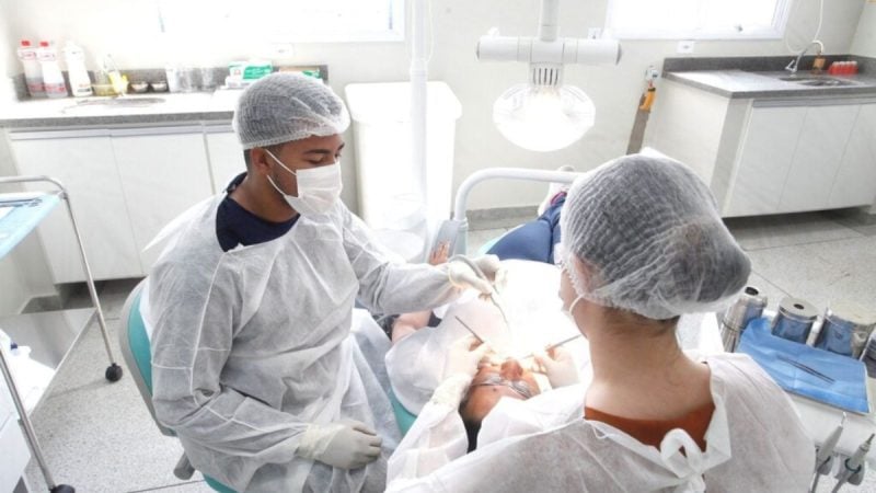 atendimento odontológico em Piracicaba: Equipe de atendimento odontológico em clínica com profissionais usando máscaras e gorros, realizando procedimento em consultório com paciente em cadeira. Piracicaba chega a 52 unidades com atendimento odontológico gratuito.