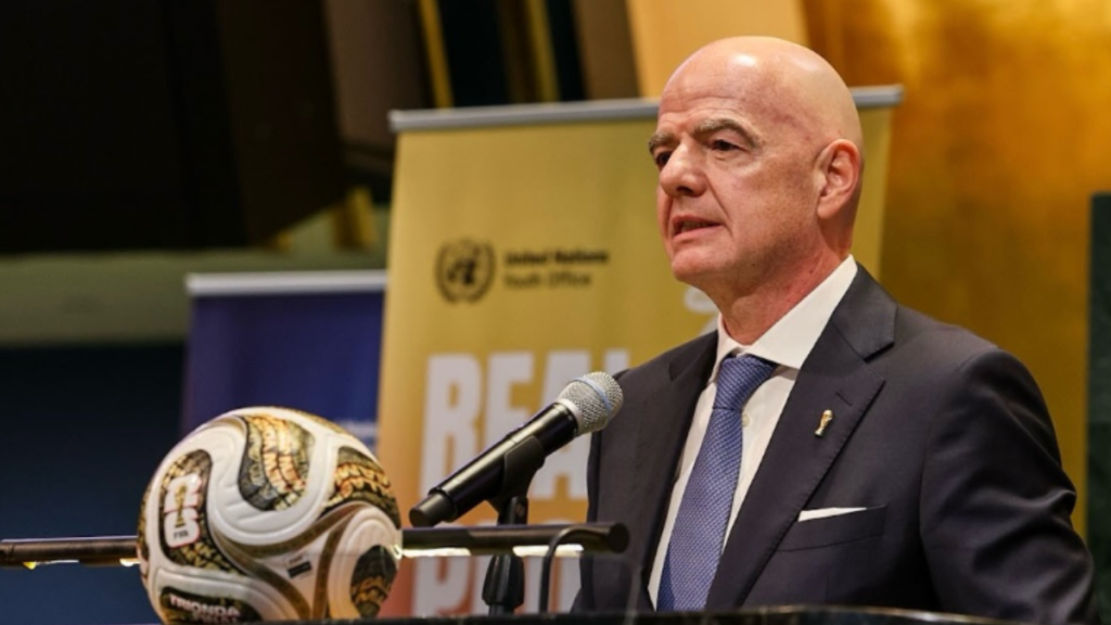 Gianni Infantino, presidente da FIFA, em foto de reunião, ao lado de microfone e bandeiras ao fundo, fazendo discurso em ambiente institucional.