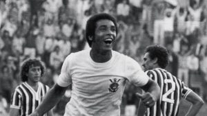 Geraldão, ídolo do Corinthians, aparece em foto em preto e branco em campo durante partida contra o Santos, marcado no centro em pose de comemoração com torcidas ao fundo.