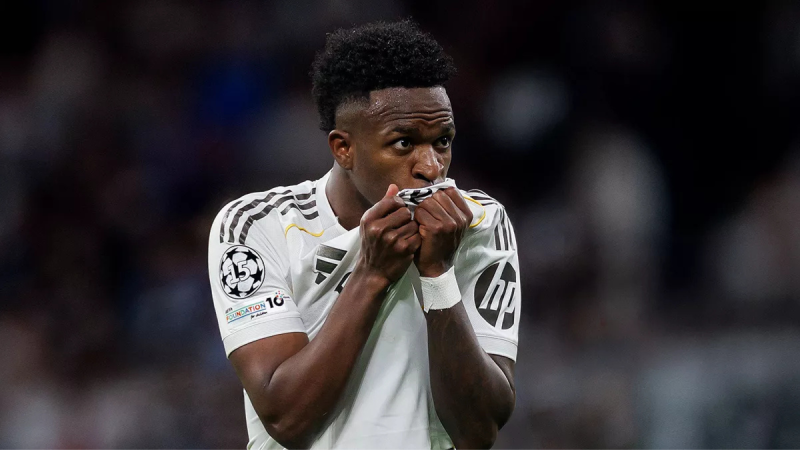 Vini Jr em partida pelo Real Madrid, segurando as mãos em frente ao rosto, com uniforme branco e escudo do clube, em foto com fundo desfocado de estádio.