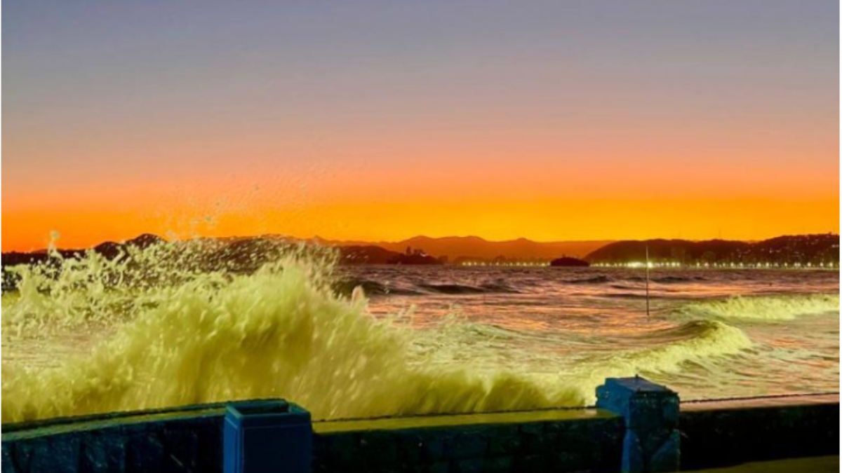 Previsão tempo baixada santista: Vista do mar agitado na Baixada Santista, com grandes ondas e espuma avançando na orla durante ventos fortes e possível ressaca ao pôr do sol.