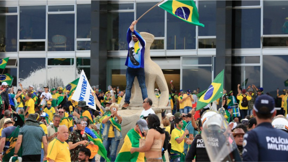Manifestantes com bandeiras do Brasil e apoio ao país em frente a um grande prédio público, em referência ao 8 de janeiro, com cartazes e clima de protesto.
