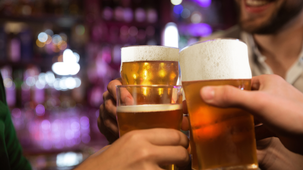 Dia Internacional da Cerveja com amigos brindando em um bar, destacando copos de cerveja dourada e clara ao fundo com luzes desfocadas, clima de comemoração.