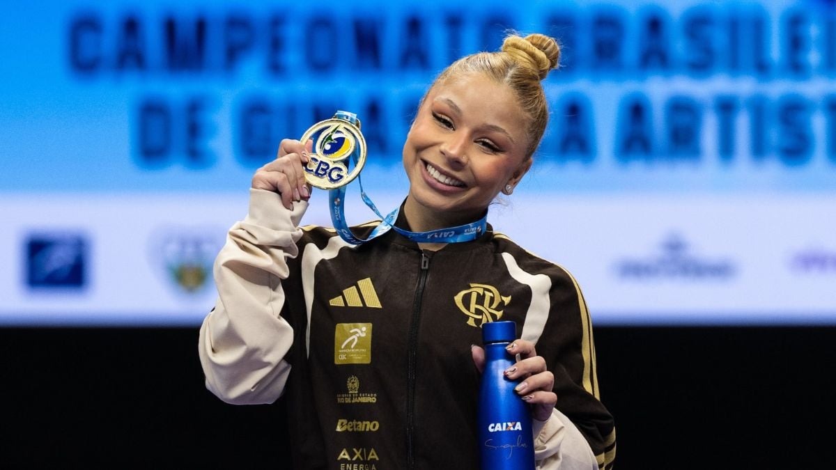 Flávia Saraiva sorrindo no pódio, exibindo dois troféus e uma garrafa azul em campeonato, com uniforma esportivo e medalha, em homenagem emocionada ao pai.