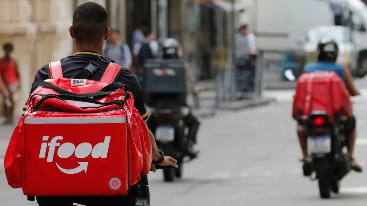 Entregador de motocicleta com bolsa vermelha iFood, em rua urbana, enquanto outros motociclistas seguem no fundo; contexto sobre medicamentos por aplicativos autorizados pela Anvisa