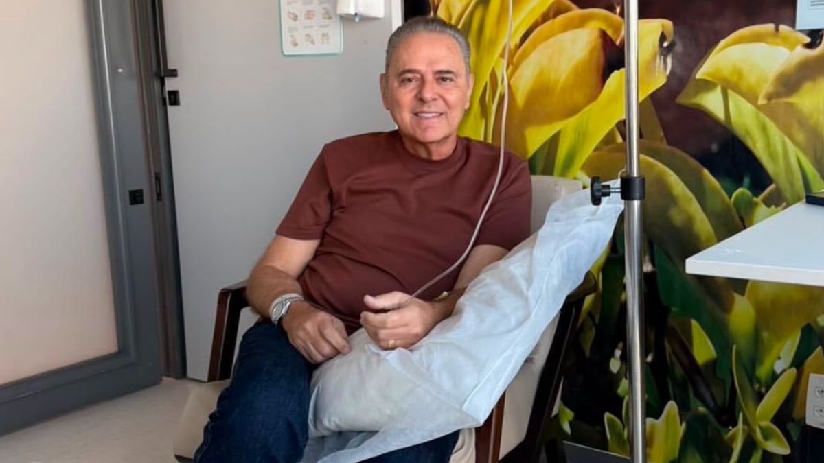 Luis Roberto sentado em uma cadeira hospitalar, com pernas cobertas por lençol branco e fones de ouvido, enquanto colegas reagem à sua volta às transmissões: “A melhor notícia do ano”.