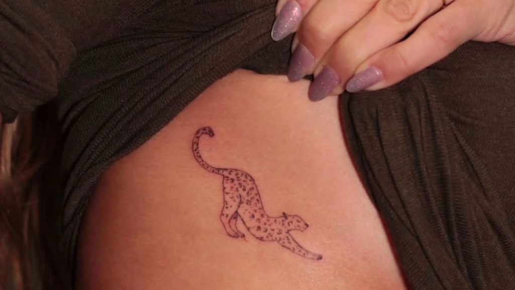 Grazi Massafera aparece em close durante sessão de tatuagem, com uma tatuagem de estilo pontilhista próxima ao seio, enquanto a mão de outra pessoa aplica a arte na pele.