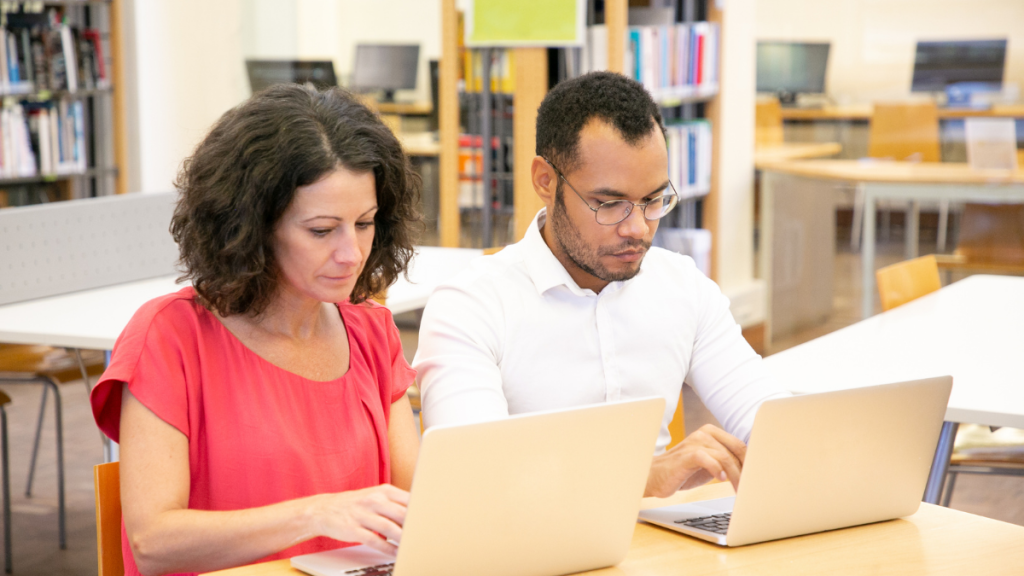 Cursos gratuitos de IA do Qualifica SP: duas pessoas estudando com notebooks em uma biblioteca, representando oportunidades de cursos gratuitos de inteligência artificial em São Paulo.
