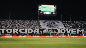 Torcida organizada do Santos em estádio lotado, com grande faixa e bandeirão do clube durante encontro com o elenco no campo