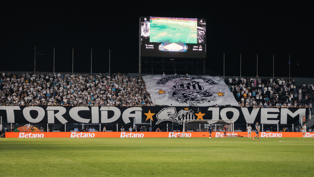 Torcida organizada do Santos em estádio lotado, com grande faixa e bandeirão do clube durante encontro com o elenco no campo