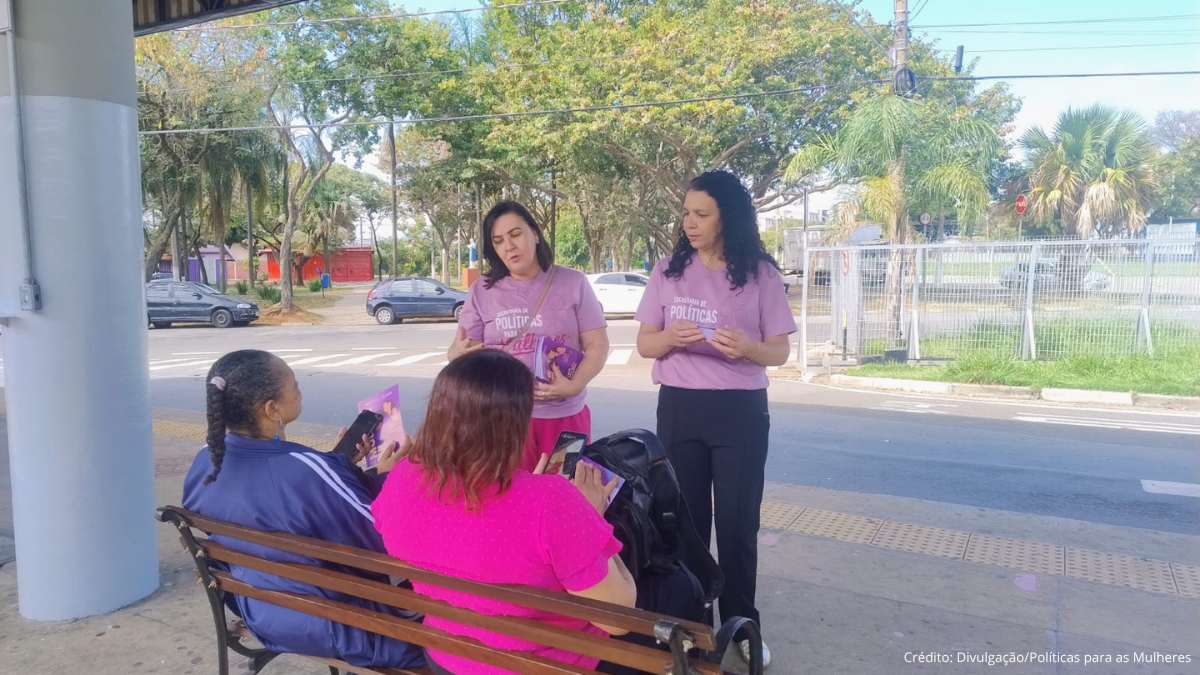 Campinas leva combate à Violência contra a mulher aos terminais de ônibus, com integrantes da ação em roupas roxas conversando com mulheres sentadas em um ponto