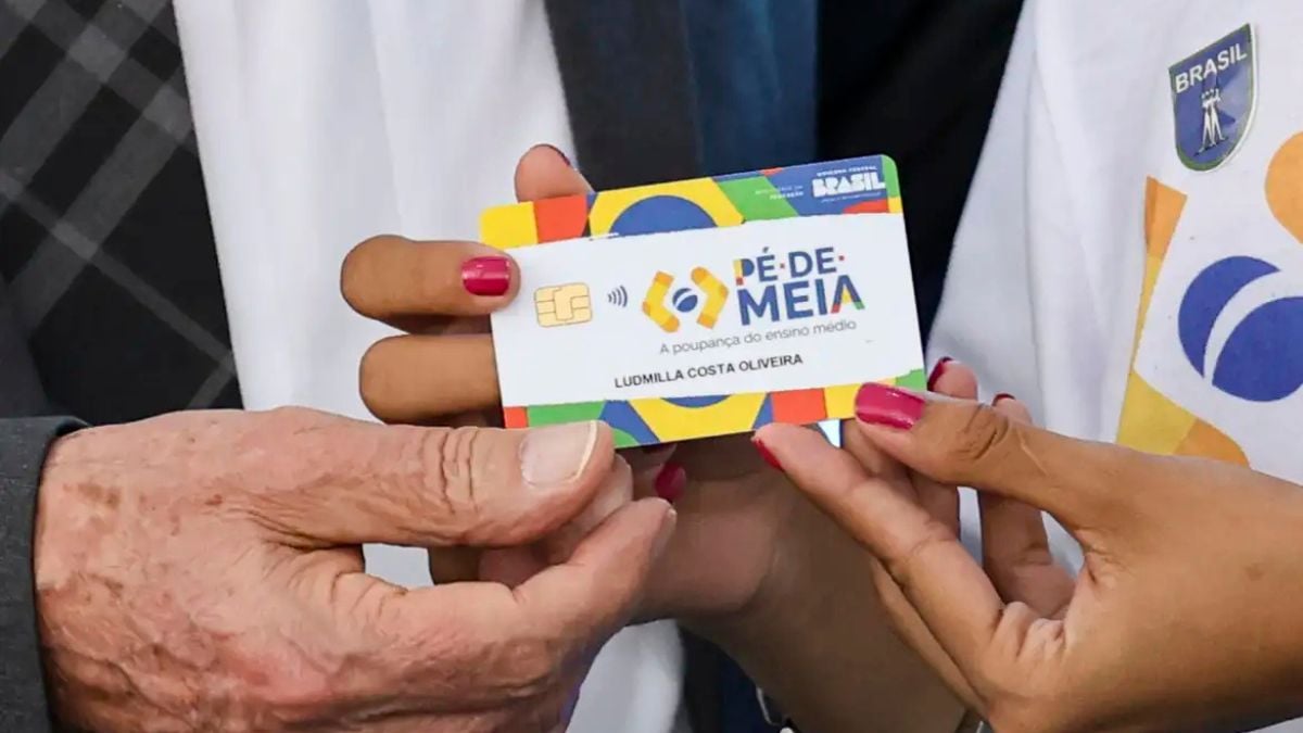Calendário pé-de-meia em agosto: mãos segurando cartão do programa Pé-de-Meia, com texto “PÉ-DE-MEI” e personagem em segundo plano, indicando quando o dinheiro cai.