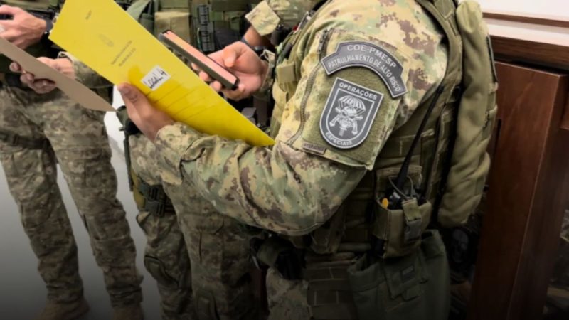 operação patrocínio Fiel: Policiais em operação na Baixada Santista, com uniformes camuflados e distintivo de operações, segurando papel e revistando documentos durante ação contra grupo suspeito de sequestrar e torturar advogado.