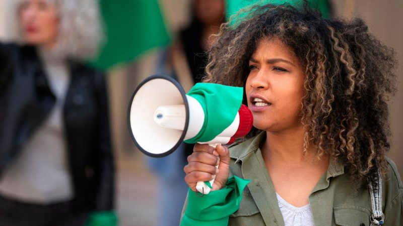Eleições 2026: mulher segura megafone verde e branco, em ambiente institucional, em referência ao início da propaganda eleitoral no domingo (16), com apelo político