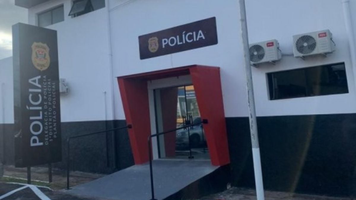 Entrada de uma delegacia com placa “POLÍCIA”, câmeras e equipamentos, em contexto de notícia sobre bebê em Sumaré desnutrido encontrado e mulher presa na cidade.