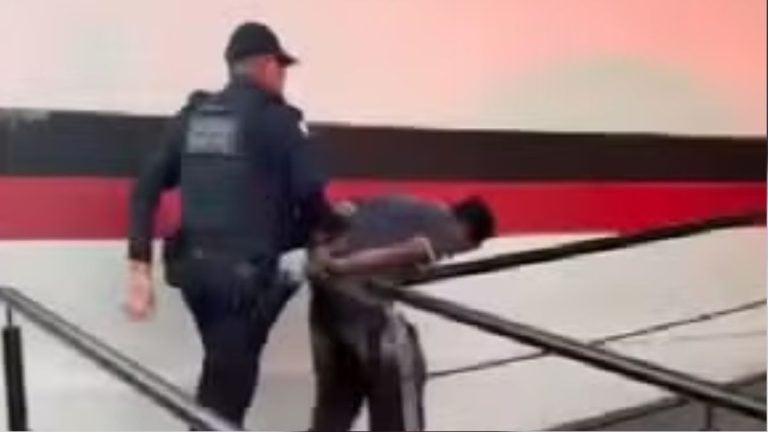 Imagem mostra um suspeito algemado escoltado por policiais na área externa, em cena relacionada a homem é solto após manter companheira em cárcere em Campinas, segundo notícia.