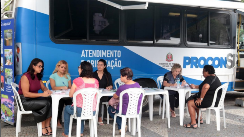Atendimento do Procon Móvel Santos em uma van azul, com equipe e moradores sentados em cadeiras plásticas durante ação de atendimento no Morro Nova Cintra.