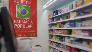 Cartaz do Governo com o logotipo “Farmácia Popular” ao lado de prateleiras com medicamentos em uma farmácia, ilustrando novas medidas do Programa Farmácia Popular.