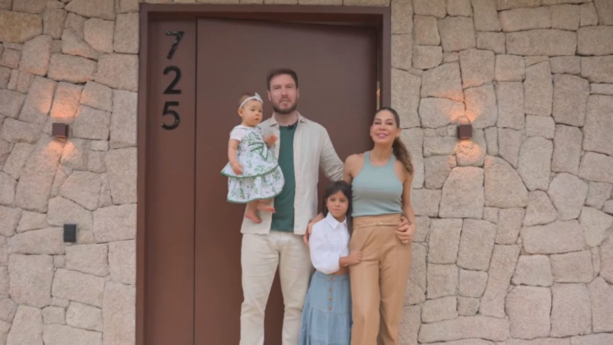 Maíra Cardi e Thiago Nigro posam em frente à entrada de uma mansão, com a família, em uma área externa de pedras e número 725 ao lado da porta