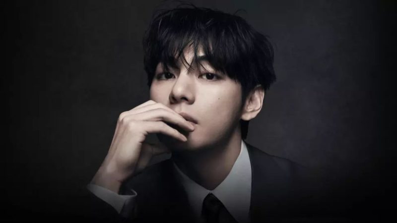 V do BTS em foto de estúdio, com cabelo escuro e expressão pensativa, apoiando o rosto na mão, em fundo escuro, em imagem de capa de notícia