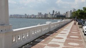 Mureta da Ponta da Praia: Vista do calçadão à beira-mar com mureta branca e faixas de piso, ao lado da água e do skyline de prédios ao fundo.
