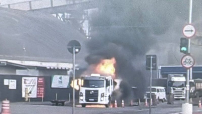 Caminhão Santos carregado com piche pega fogo na via, com chamas saindo da parte frontal e fumaça ao redor, em Santos, e deixa motorista ferido.